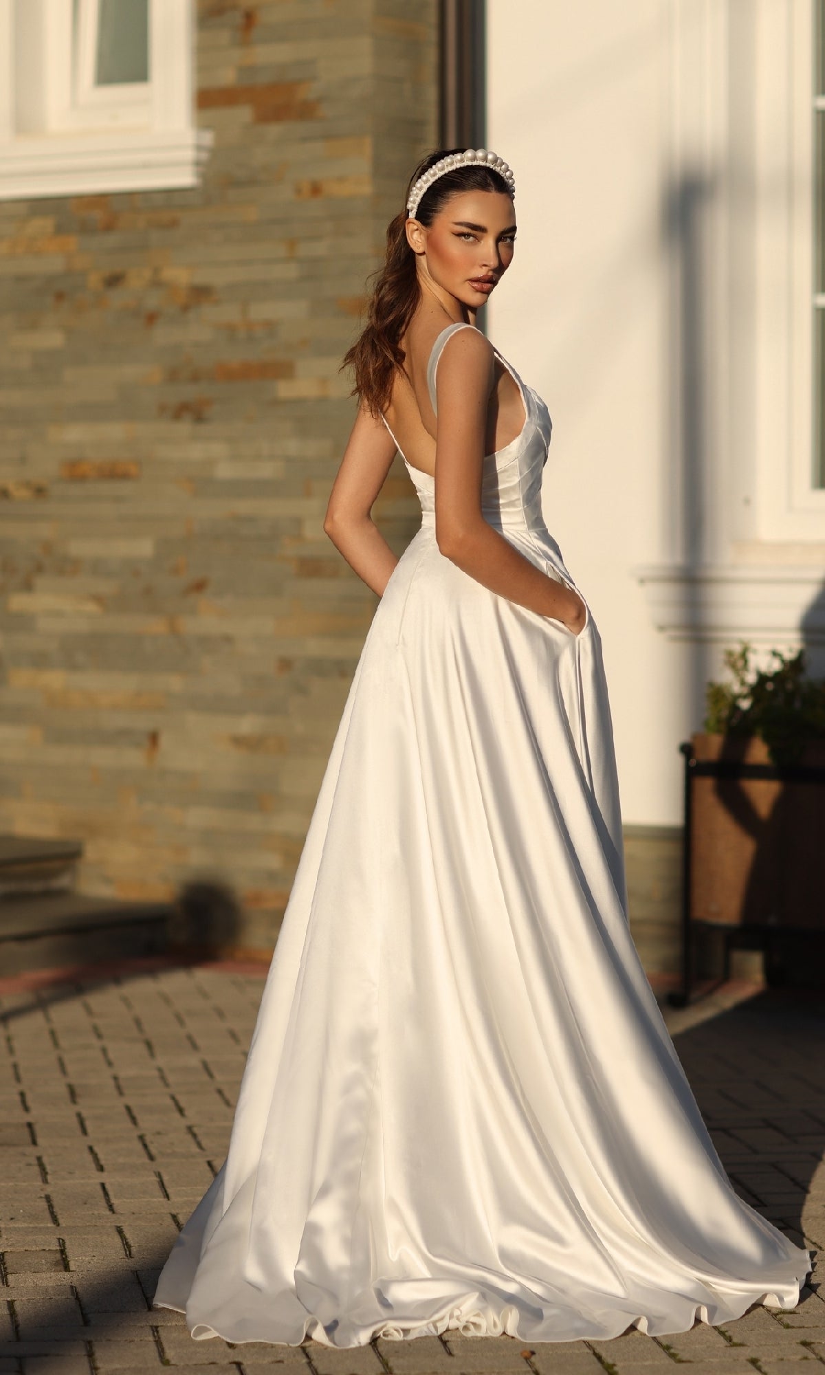 A woman in the Off-White Satin Prom Ball Gown: Tina Holly BB210W, featuring an a-line silhouette and pockets, stands on a stone walkway outdoors, glancing over her shoulder.